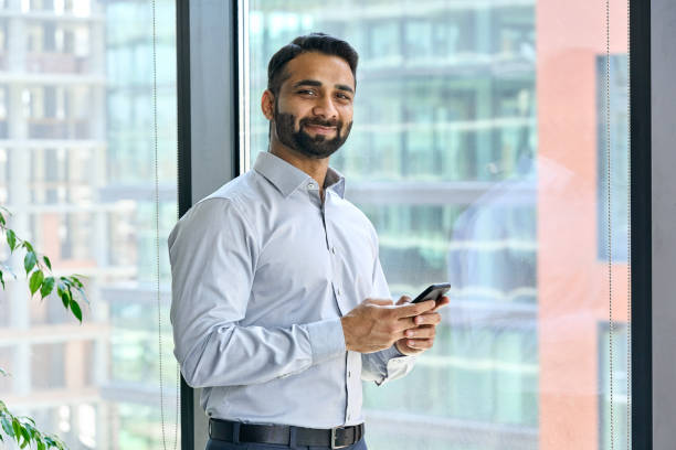 istock Smiling indian businessman holding phone standing in office at panoramic window. 1334472373