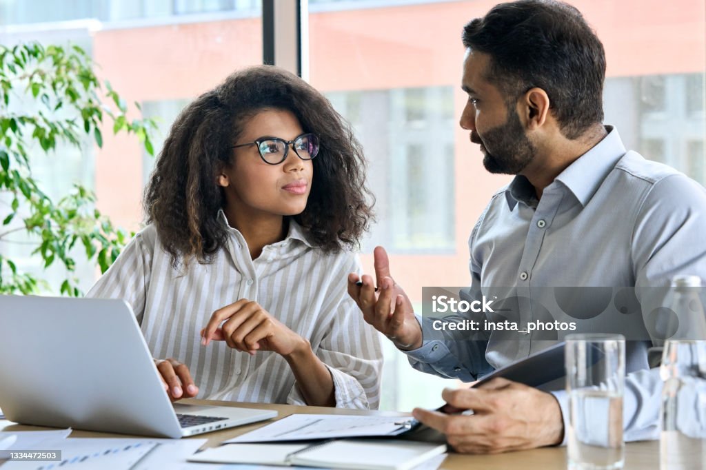 Indian ceo mentor leader talking to female trainee using laptop at meeting. Businessman indian executive manager ceo talking to female African American coworker, using laptop. Diverse multicultural professional partners group discussing business plan at board room meeting. Advice Stock Photo