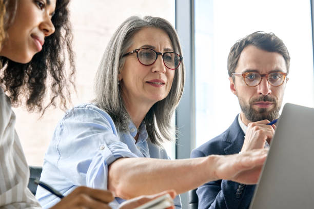 Diverse business executive team discuss corporate report using laptop at table.