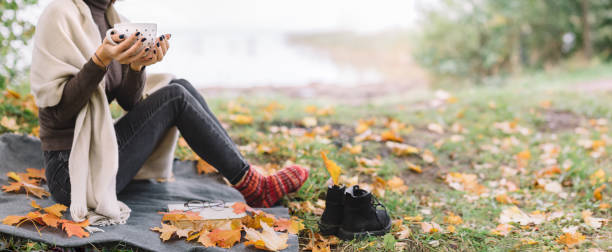 Slim girl sitting on a plaid and drinking coffee or tea from white polka dot cup. Colorful maple leaves around. Warming beverage for autumn season. Copy space.
