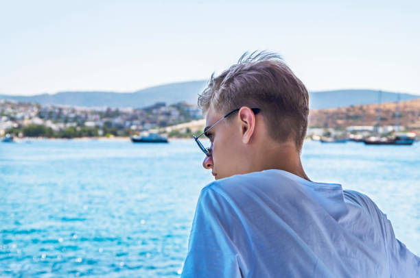 giovane, adolescente di età tra i 15 e i 18 anni appoggiato sulla spiaggia con occhiali da sole, godendosi la vista su uno sfondo blu di acqua dell'oceano - teenager 14 15 years 13 14 years cheerful foto e immagini stock