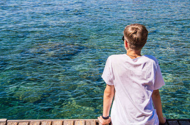 giovane, adolescente di età tra i 15 e i 18 anni appoggiato sulla spiaggia con occhiali da sole, godendosi la vista su uno sfondo blu di acqua dell'oceano - teenager 14 15 years 13 14 years cheerful foto e immagini stock