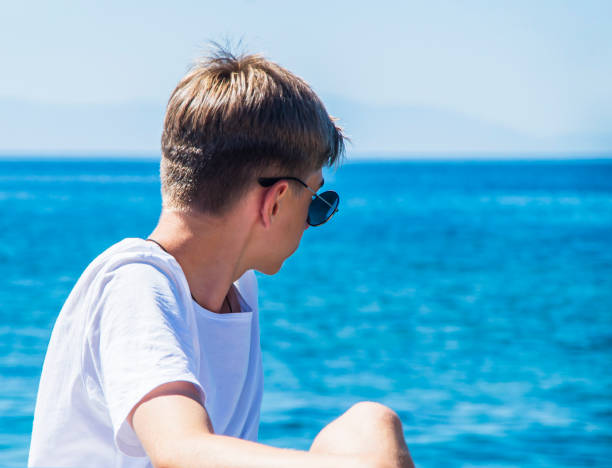 giovane, adolescente di età tra i 15 e i 18 anni appoggiato sulla spiaggia con occhiali da sole, godendosi la vista su uno sfondo blu di acqua dell'oceano - teenager 14 15 years 13 14 years cheerful foto e immagini stock