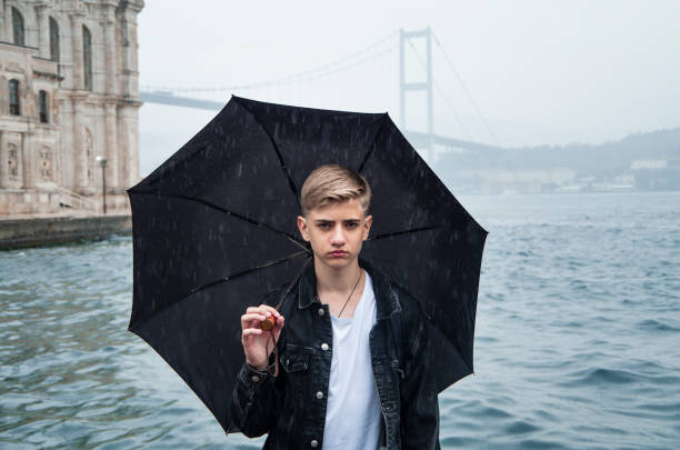 regen, schlechtes wetter. gutaussehender ernsthafter teenager 15-18 jahre alt, der einen regenschirm hält, während er vor der kulisse der ortakoy-moschee und der bosporus-brücke in istanbul türkei spazieren geht.weiche selektive fokussierung. - 12 13 years stock-fotos und bilder