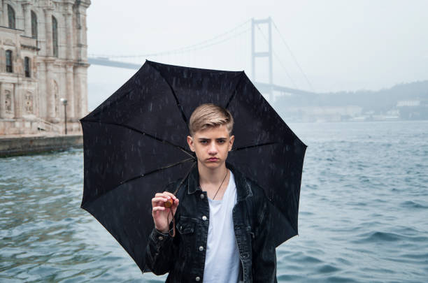 雨、悪天候。イスタンブールトルコのオルタコイモスクとボスポラス橋を背景に歩いている間、傘を持っているハンサムな深刻なティーンエイジャー15-18歳の男。 - 12 13 years ストックフォトと画像