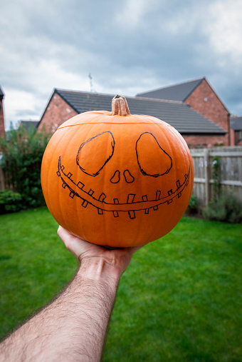 A point of view shot of an unrecognisable person holding a pumpkin which has a spooky face design drawn on it, ready to be carved out.