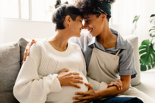 Married lesbian couple expecting a baby. Pregnant lesbian couple smiling and embracing each other while sitting in their living room. Expectant female couple bonding fondly at home.