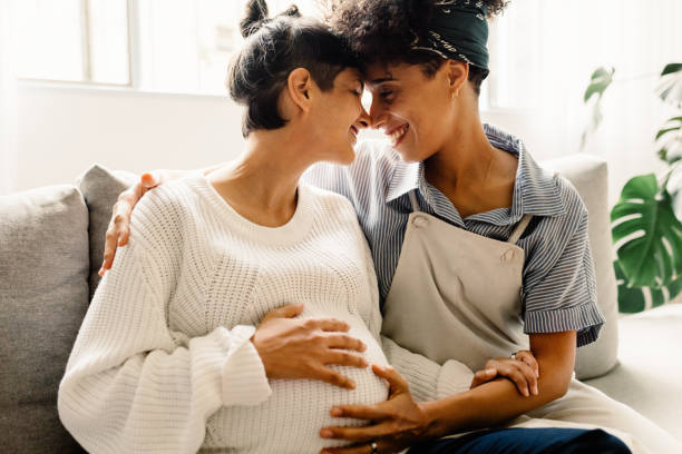 Married lesbian couple expecting a baby Married lesbian couple expecting a baby. Pregnant lesbian couple smiling and embracing each other while sitting in their living room. Expectant female couple bonding fondly at home. lgbtqia people stock pictures, royalty-free photos & images