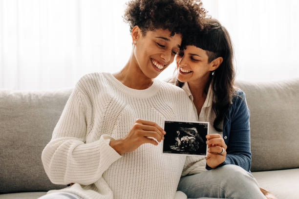 Same-sex pregnant couple holding up their ultrasound scan Same-sex pregnant couple holding up their ultrasound scan. Expectant lesbian couple smiling cheerfully while holding an ultrasound picture of their unborn baby. Young queer couple expecting a baby. lgbtqia rights photos stock pictures, royalty-free photos & images