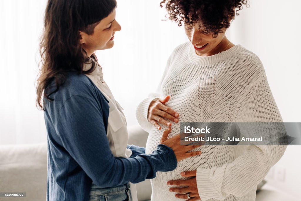 Happy woman touching a surrogate mother's belly bump Happy young woman touching a surrogate mother's belly bump. Woman smiling while feeling the movement of a pregnant woman's baby. Young woman spending time with her surrogate at home. Pregnant Stock Photo