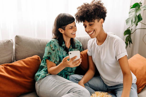 Two lesbian lovers using a smartphone together at home Two lesbian lovers using a smartphone together at home. Lesbian couple smiling cheerfully while surfing the net on a smartphone. Young LGBTQ+ couple sitting together in their living room. gay couple photos stock pictures, royalty-free photos & images