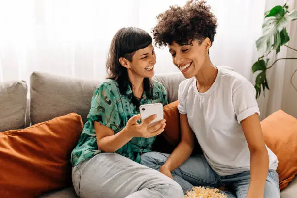 Two lesbian lovers using a smartphone together at home. Lesbian couple smiling cheerfully while surfing the net on a smartphone. Young LGBTQ+ couple sitting together in their living room.