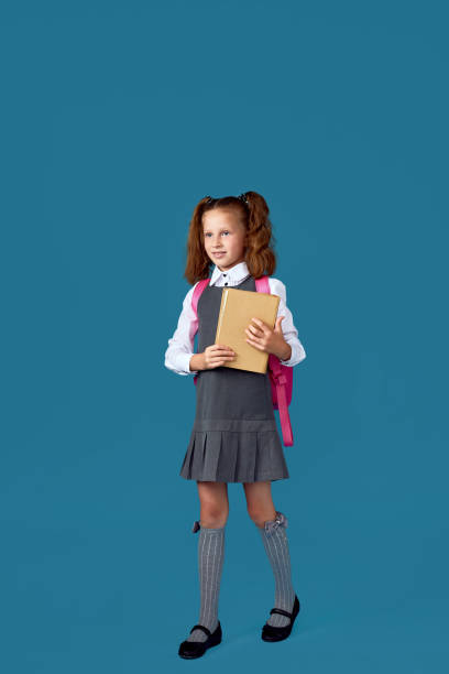 istock cute schoolgirl with backpack and book full length. 1334488526