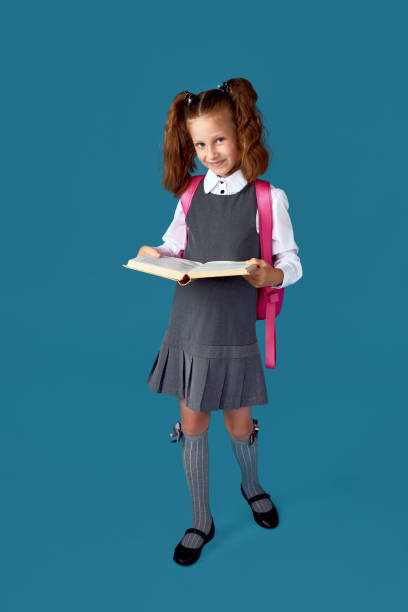 istock cute schoolgirl with backpack and book full length. 1334488605