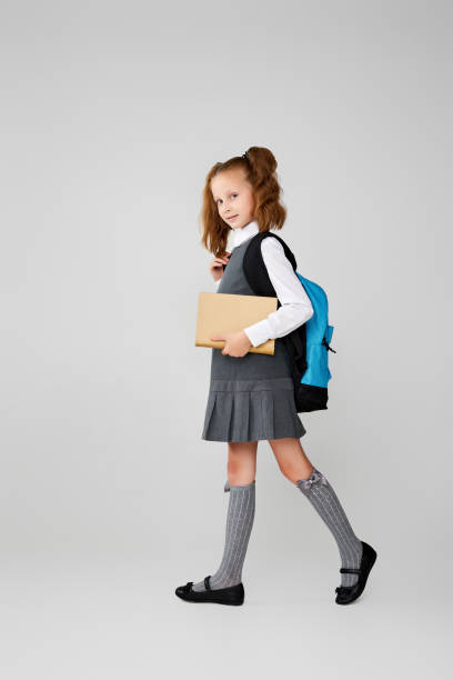istock cute schoolgirl with backpack and book full length. 1334489730