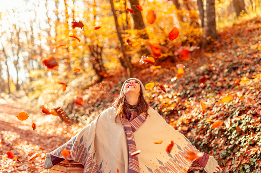 Cheerful young woman having fun spending sunny autumn day in nature, looking up with arms spread, watching the colorful leaves fall on the forest path