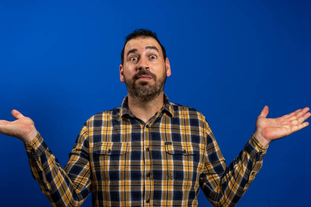 waist up shot of clueless brunet european man shrugs shoulders raises palms feels doubtful dressed in casual yellow plaid shirt isolated over blue background. people hesitation and puzzlement concept - blank expression head and shoulders horizontal studio shot imagens e fotografias de stock