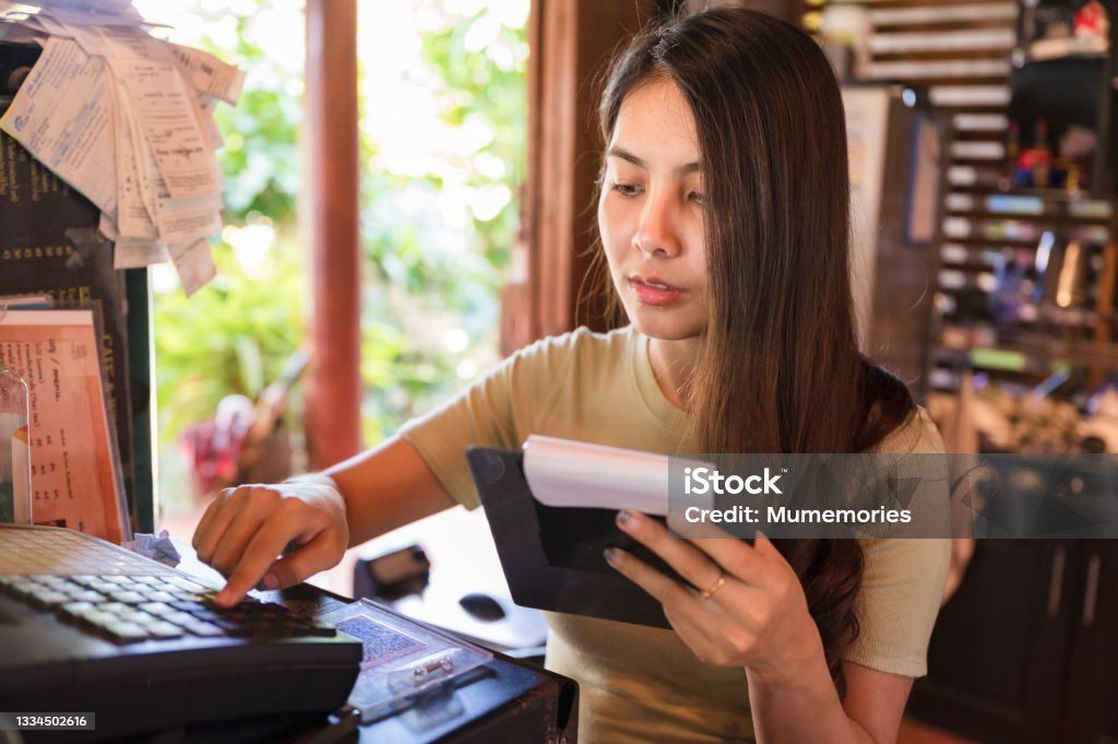 Asian woman cashier checking order customer and pressing cash register at counter in restaurant Young asian woman cashier checking order customer and pressing cash register at wooden counter in restaurant Business Stock Photo