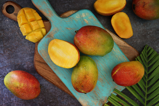 Image of wooden, turquoise chopping board, pile of ripe, tropical mango fruit, red skins on wood grain surface against black background, surrounded by palm leaf fronds, one cut into hedgehog cube shapes exposing the orange flesh, elevated view
