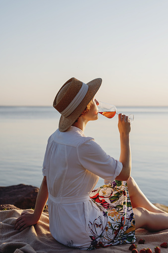 Young woman in straw hat and summer dress drinking wine at the sea shore on sunset.