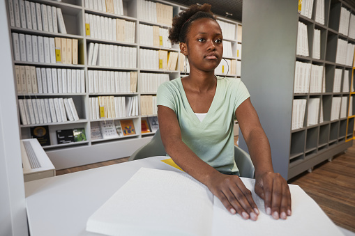 Portrait of young African-American woman reading Braille book in college library, copy space