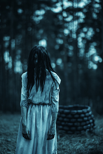 Girl with long black hair in image of scary ghost zombie walks among dark forest against background of stone well.