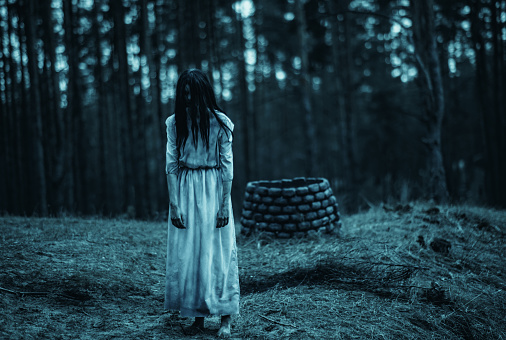Girl with long black hair in image of scary ghost zombie walks among dark forest against background of stone well.
