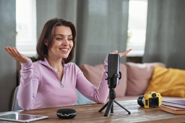 istock Cheerful young Caucasian female blogger sitting at table and gesturing hands while shooting for social media using smartphone on tripod 1334512478
