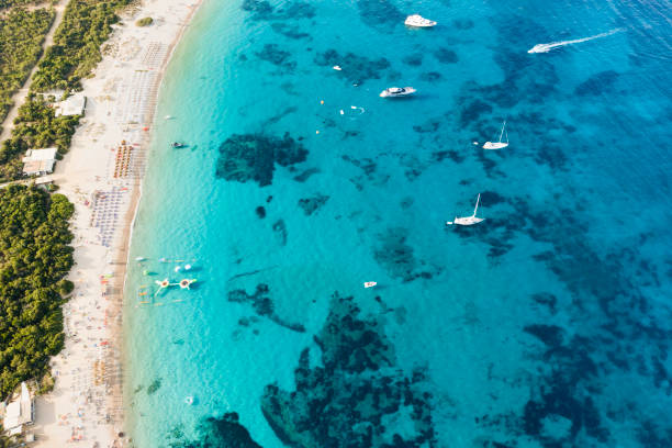 View from above, stunning aerial view of a green coastline with a white sand beach and and boats sailing on a turquoise water at sunset. Cala di volpe beach, Costa Smeralda, Sardinia, Italy. View from above, stunning aerial view of a green coastline with a white sand beach and and boats sailing on a turquoise water at sunset. Cala di volpe beach, Costa Smeralda, Sardinia, Italy. Cala Di Volpe stock pictures, royalty-free photos & images