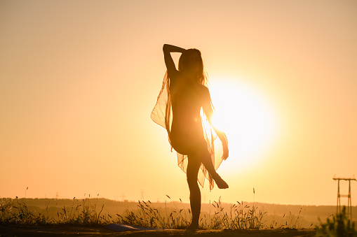 Beautiful fit girl exercise, dancing in transparent tunic and warm sun light with water and warm sky view