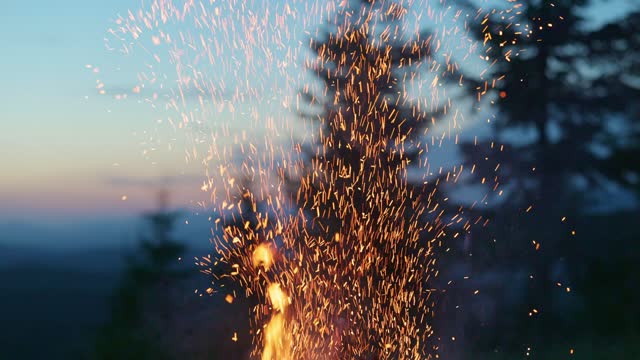 Bright sparks fly from the fire and float in the air. Bonfire, wild camping, hiking.  Mountains, trees and sunset sky on the background. Slow motion, 4K