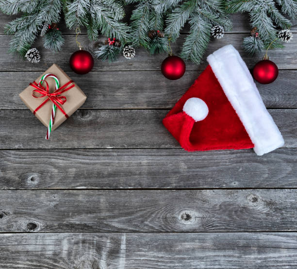 istock Snow covered evergreen branches plus bright red ornaments, Santa cap and gift box on faded wooden planks for a merry Christmas or happy New Year holiday celebration concept 1334532815