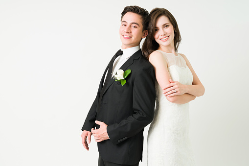 Cheerful bride in a wedding gown and handsome groom smiling while making eye contact in front of a studio background