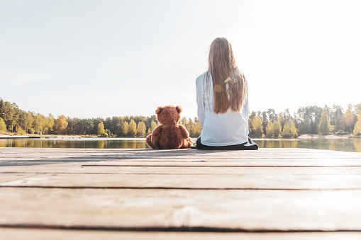 Little girl with stuffed toy teddy bear sitting on wooden pier near calm lake