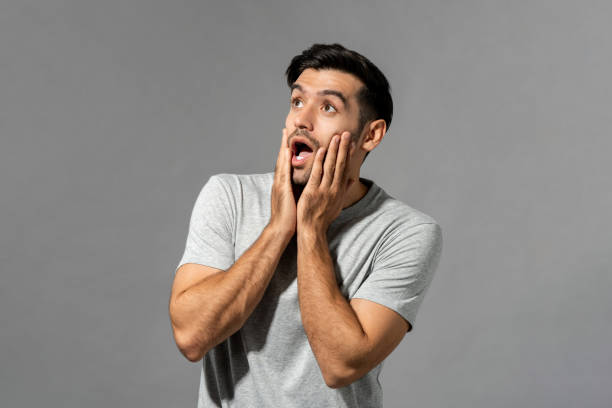 istock Shocked young Caucasian man with hands on cheeks gasping and looking up in light gray isolated studio background 1334547875