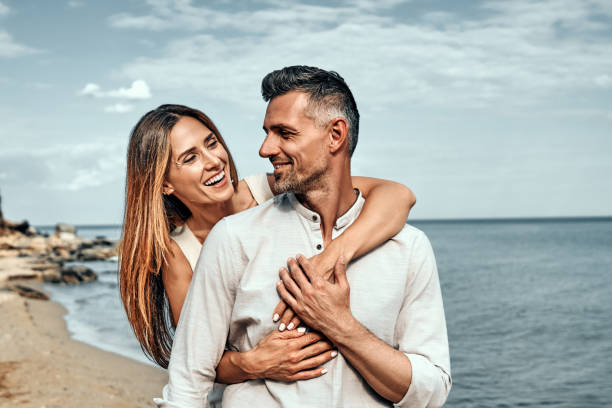 mujer sosteniendo a su marido por detrás de pie en una playa en un día soleado. - standing smiling two people 30s fotografías e imágenes de stock