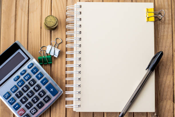 istock Plain Spiral Notebook With Pen Beside Calculator And Coins On Table. Simple Notepad And Ballpen Alongside Calculation Machine Placed On Top Of Desk. 1334548409