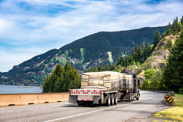 istock Black classic big rig semi truck transporting covered cargo on flat bed semi trailer running on the winding highway road along the river and mountain 1334554985