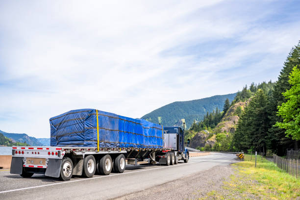 istock Dark blue classic big rig semi truck transporting covered cargo on flat bed semi trailer moving on the road along the river 1334556432
