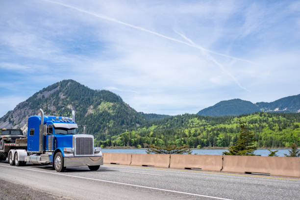 istock Bright blue big rig classic semi truck tractor with chrome accessories and sleeping cab compartment transporting car on step down semi trailer running on the road along river in Columbia Gorge area 1334558379