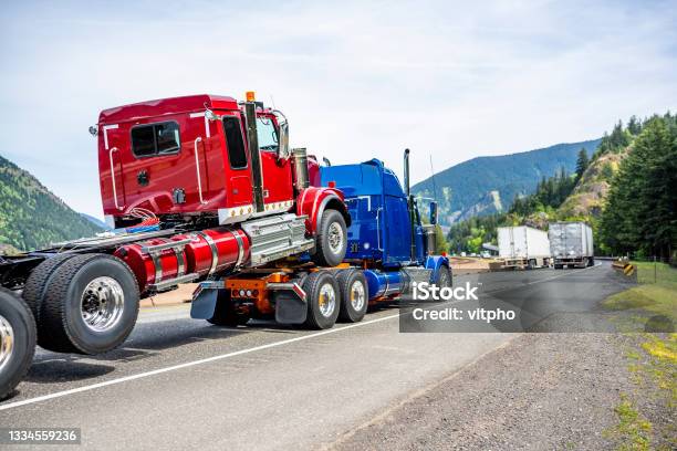 Powerful Blue Big Rig Semi Truck Transporting Another Semi Truck Tractor Immersed By The Front Wheels On The Frame Of The First Truck Running On The Road Along The River Stock Photo - Download Image Now