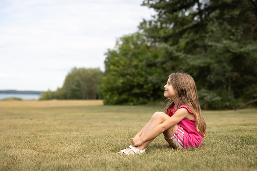 Young girl sitting on the grass in profile with knees up smiling and looking up towards sky with trees and a lake in the background in the summer with copy space