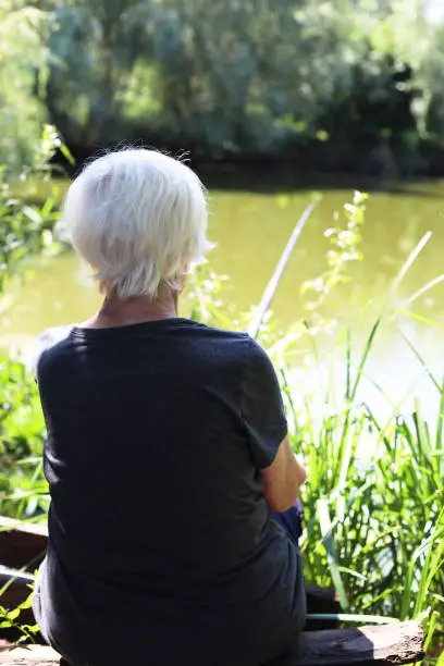 Photo of An elderly gray-haired woman is fishing on the lake. View from the back, holding a fishing rod in his hands.