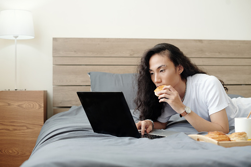 Handsome young man with long hair lying on bed, eating fresh bun and watching webinar or tutorial on laptop