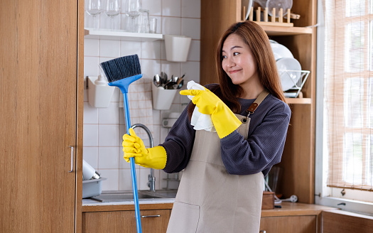 A happy housewife wearing protective glove, holding and pointing finger at a broom while cleaning in the kitchen at home