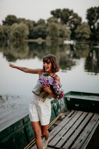 Attractive cheerful female standing on the rustic boat tied to the shore of the lake, balancing and holding a lot of fresh colorful flowers.