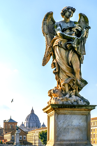 A warm sunset light envelops the statue of an Angel on the balustrade of Ponte Sant'Angelo, along the Tiber River, in the historic heart of Rome. This bridge was built under the emperor Hadrian in 134 AD. to connect the imperial city with his mausoleum, currently Castel Sant'Angelo. Centuries later, under the pontificate of Pope Clement IX, the current balustrade was built based on a project by Giovanni Lorenzo Bernini, on which ten statues of the Angels of the Passion of Christ were placed, made in 1669 by the pupils of Bernini himself. In the background, the profile of the dome of St. Peter's Basilica. In 1980 the historic center of Rome was declared a World Heritage Site by Unesco. Image in high definition format.