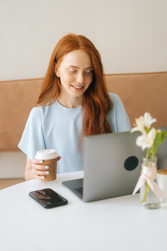 Vertical portrait of happy young beautiful woman typing on laptop and drinking coffee from cup while working remotely at cozy cafe. Pretty redhead Caucasian lady remote working or studying.