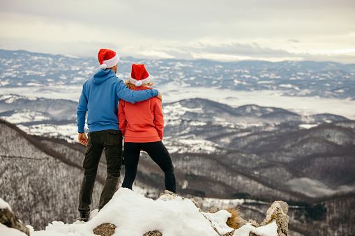 Mid adult couple standing on viewpoint and enjoying while watching mountain landscape at winter. He is embracing her and they boat wear Santa Claus hats