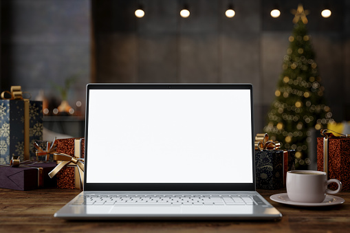 Blank Screen Laptop, Gift Boxes And Coffee Cup On The Table. Blurred Living Room With Christmas Tree Background